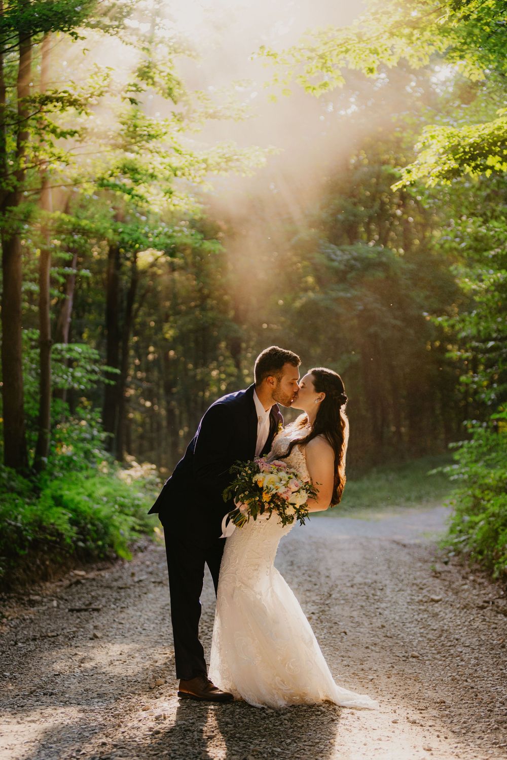 Bride and groom share a magical kiss on a sunlit forest path with golden rays.