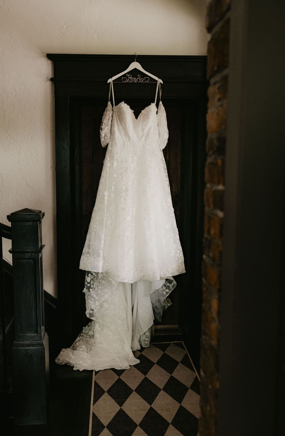 Elegant off-shoulder lace wedding gown hanging on a personalized hanger in a doorway.
