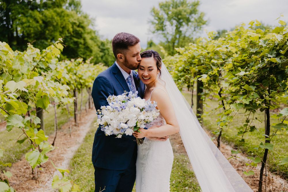 Bride and groom share a joyful embrace in a vineyard holding a white floral bouquet.