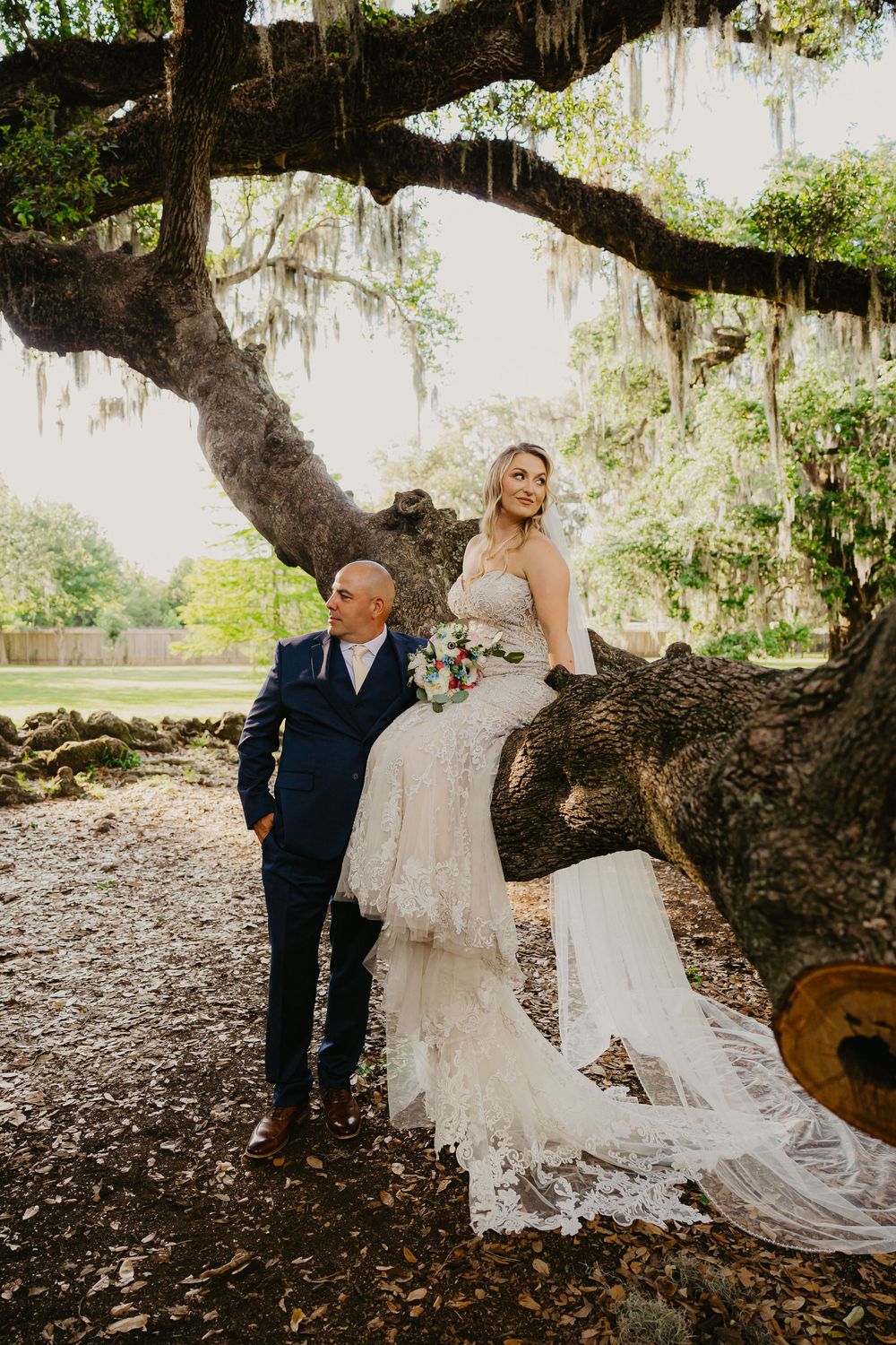 Blonde bride in lace gown sits on a mossy oak tree with groom beside her.