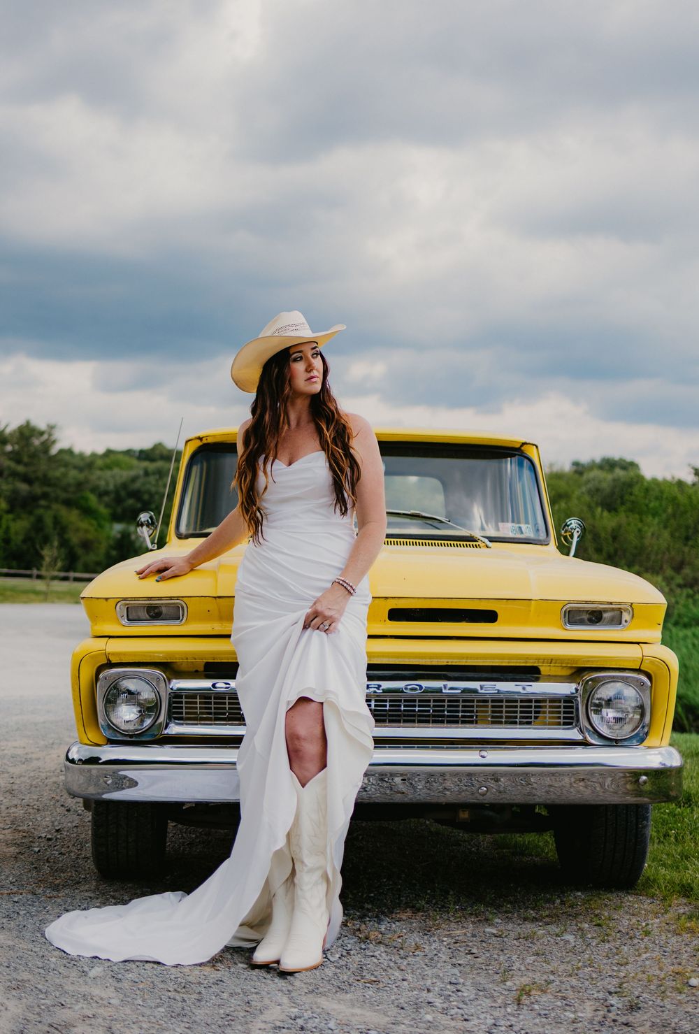 Bride in white gown, cowboy hat, and white boots leaning against a vintage yellow Chevy truck on a gravel road.