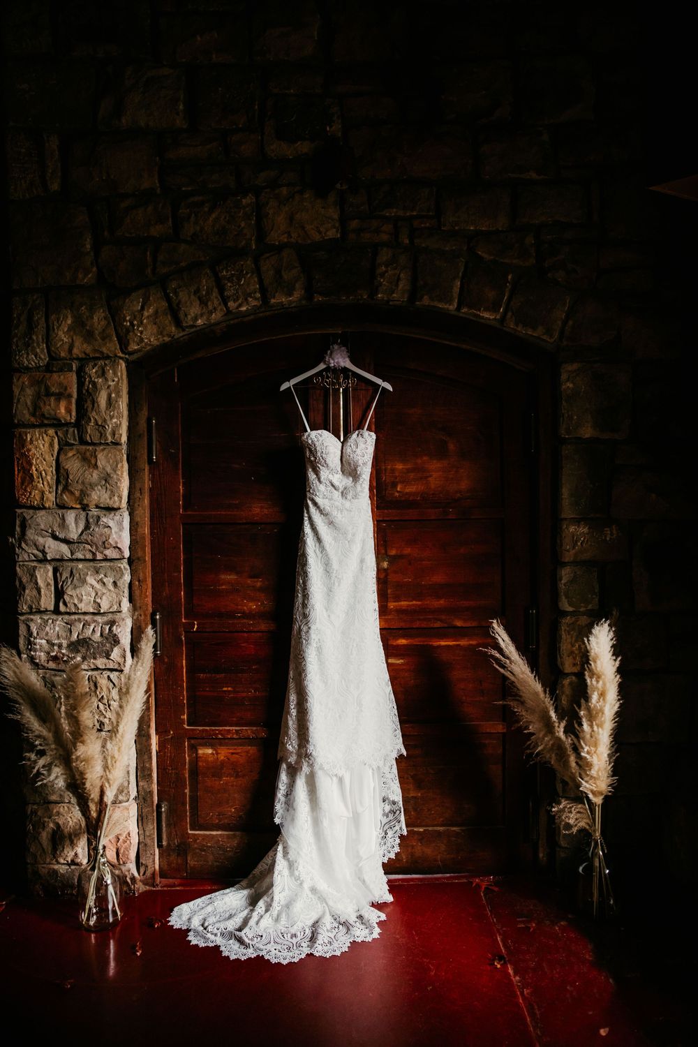 Strapless lace wedding gown hanging on wooden doors framed by stone archway and pampas grass.