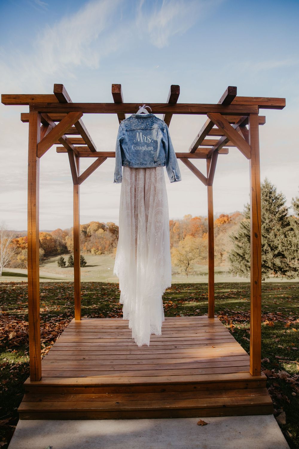 A custom denim jacket reading Mrs. hangs with a white tulle wedding dress on a wooden pergola arch outdoors in autumn.