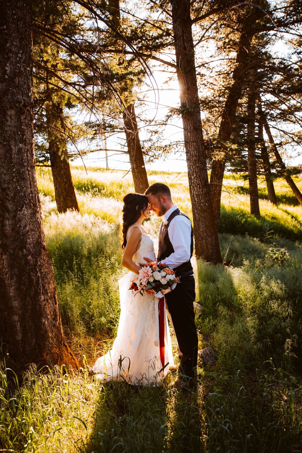 Bride and groom share a romantic moment among tall pine trees in golden sunset light.