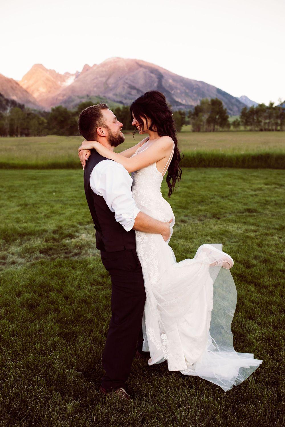 Groom lifting bride in white gown outdoors with mountain backdrop at sunset during romantic wedding photo.