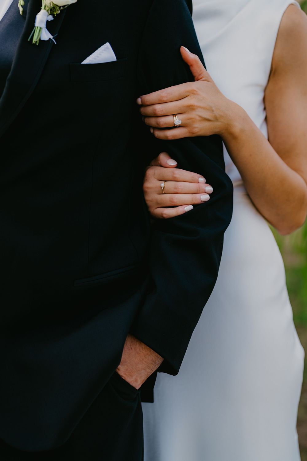 Close-up of bride and groom's hands showing wedding and engagement rings on black suit.