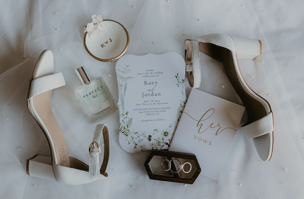 Elegant wedding flat lay featuring white heels, floral invitations, perfume, vow books, and gold rings on white fabric.