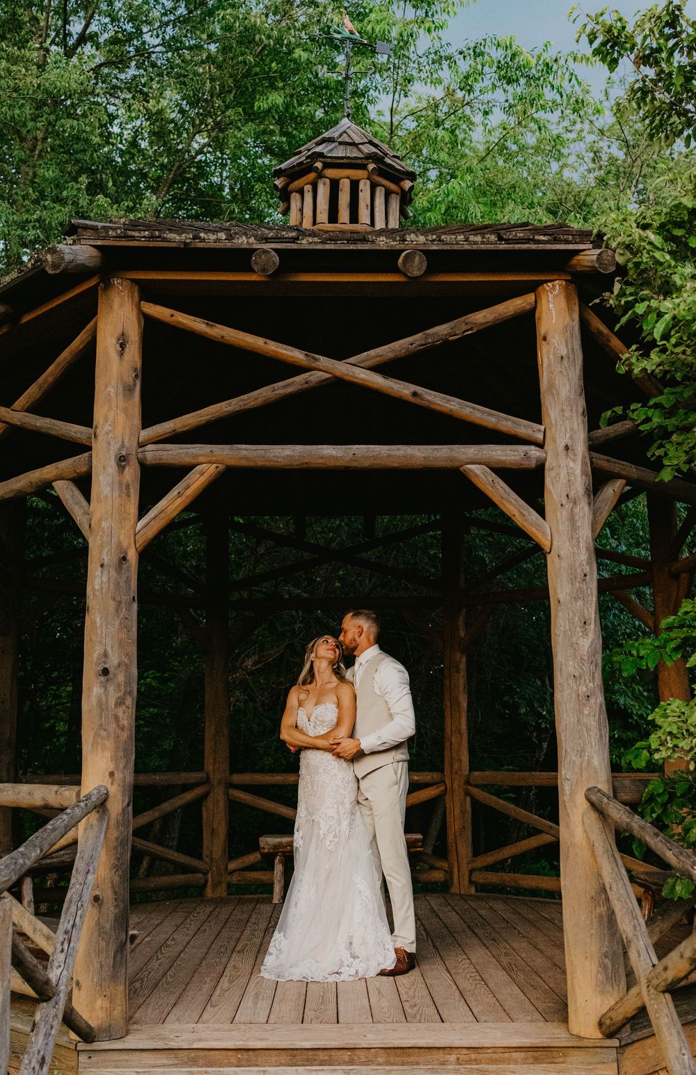 A bride and groom share a romantic moment under a rustic wooden gazebo surrounded by lush green trees.