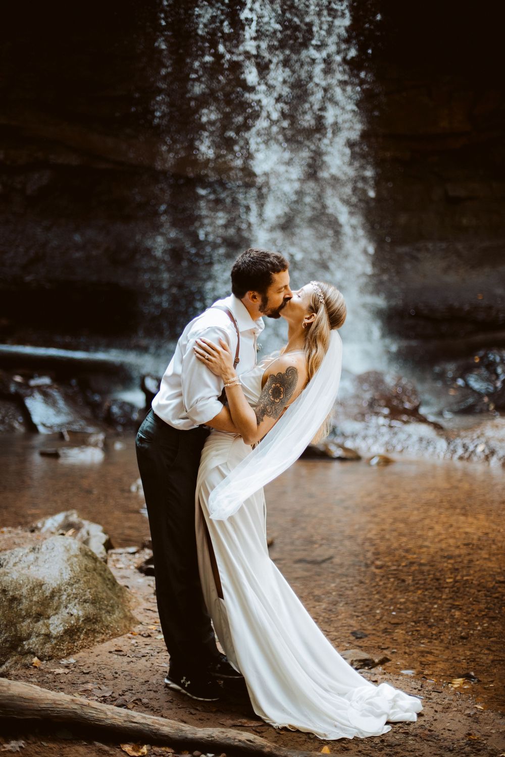 Bride and groom sharing a romantic kiss in front of a waterfall, bride in flowing white gown.