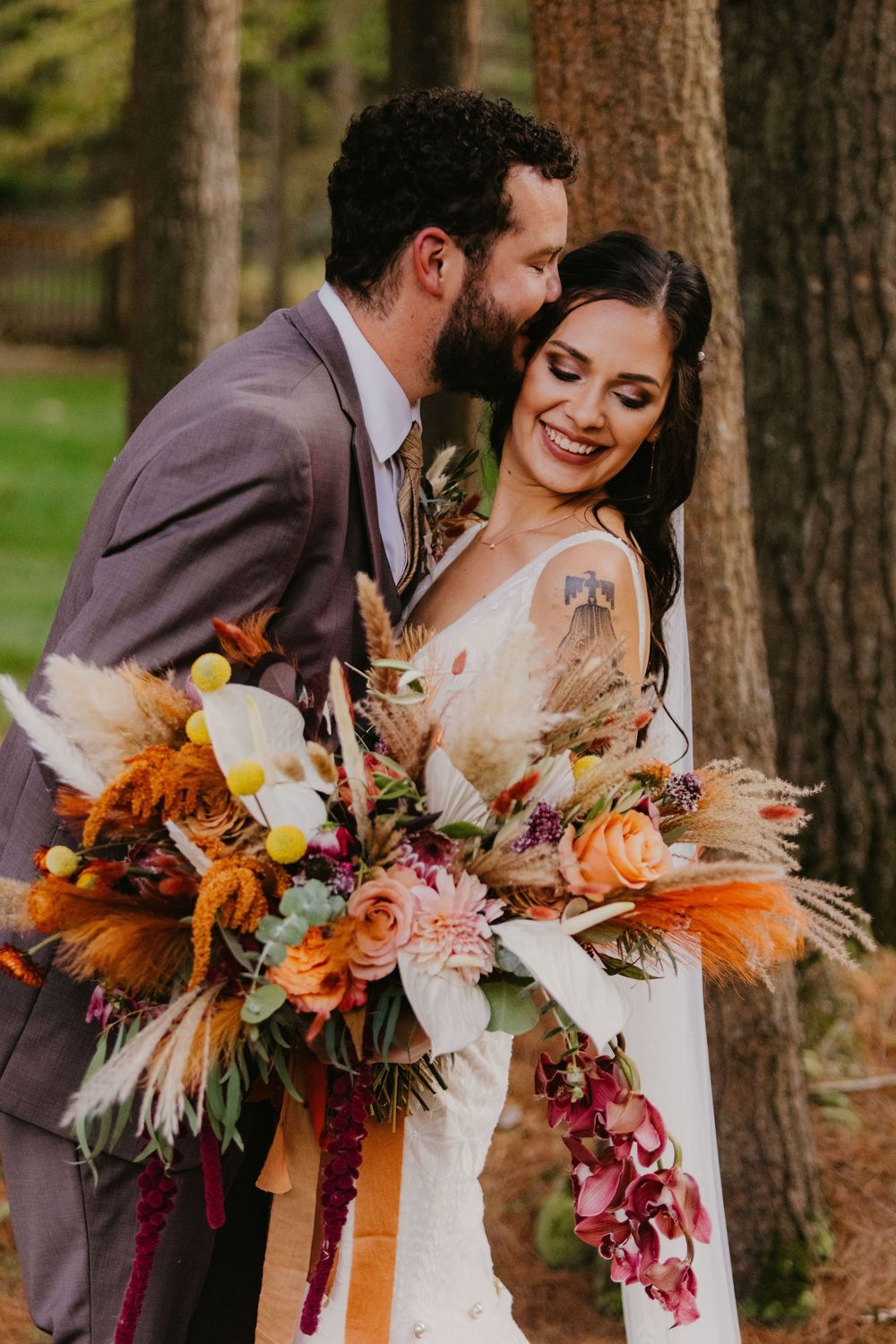 Bride smiles holding colorful boho bouquet with pampas grass as groom kisses her cheek outdoors.