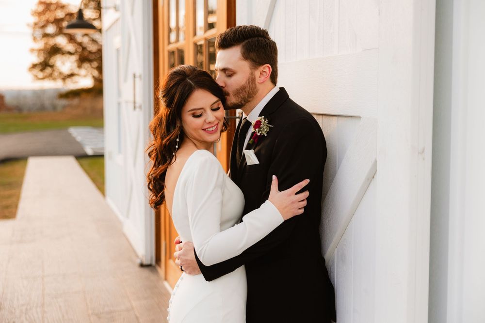 Bride in white gown embraces groom in black tuxedo against white barn wall at golden hour wedding.