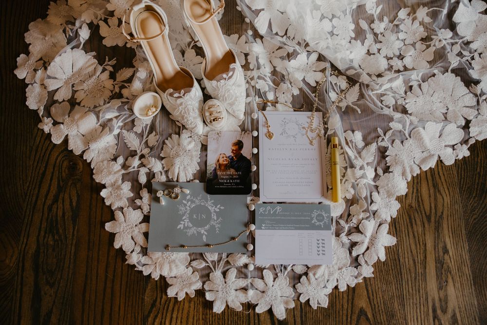 Flat lay of wedding details including floral heels, invitation suite, and lace bridal gown on wooden floor.