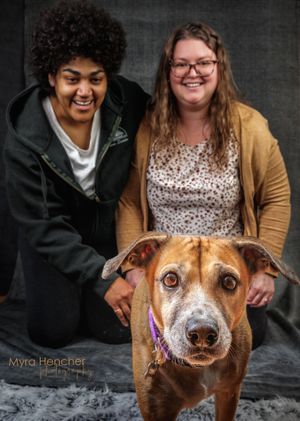 Two people smiling with a brown dog in a professional portrait photo by Myra Hencher Photography.