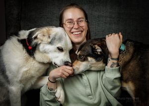 Smiling young woman with glasses hugging two dogs, a husky and a small mixed breed, in a cozy indoor setting.