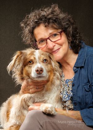 A smiling woman with curly dark hair and red glasses holds a fluffy brown and white Australian Shepherd dog in a studio portrait.