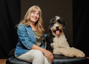 Smiling blonde woman in denim jacket poses with a fluffy black and white Bernedoodle dog on a dark studio background.