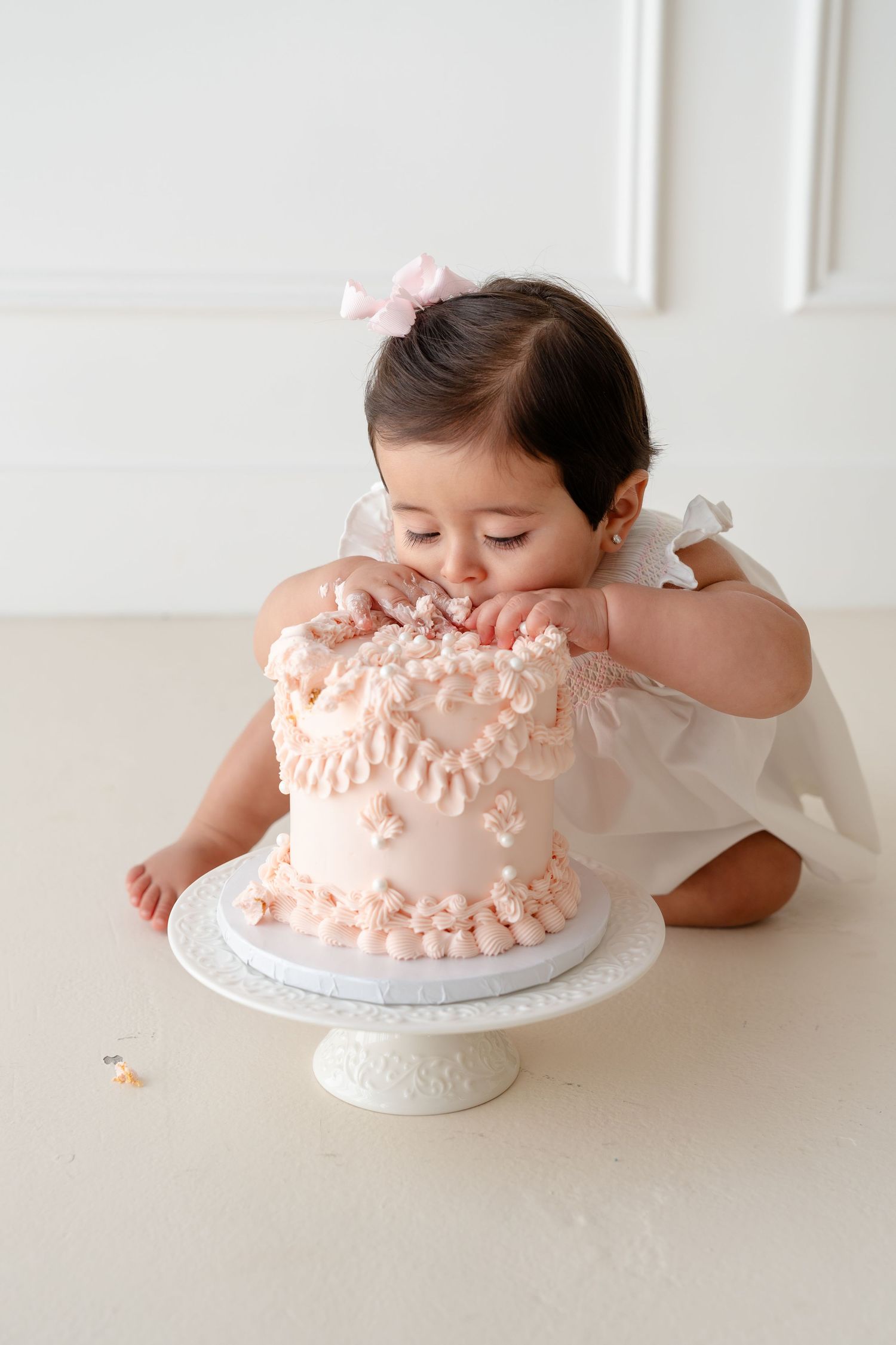 Baby girl in white dress eating pink first birthday smash cake on white cake stand.