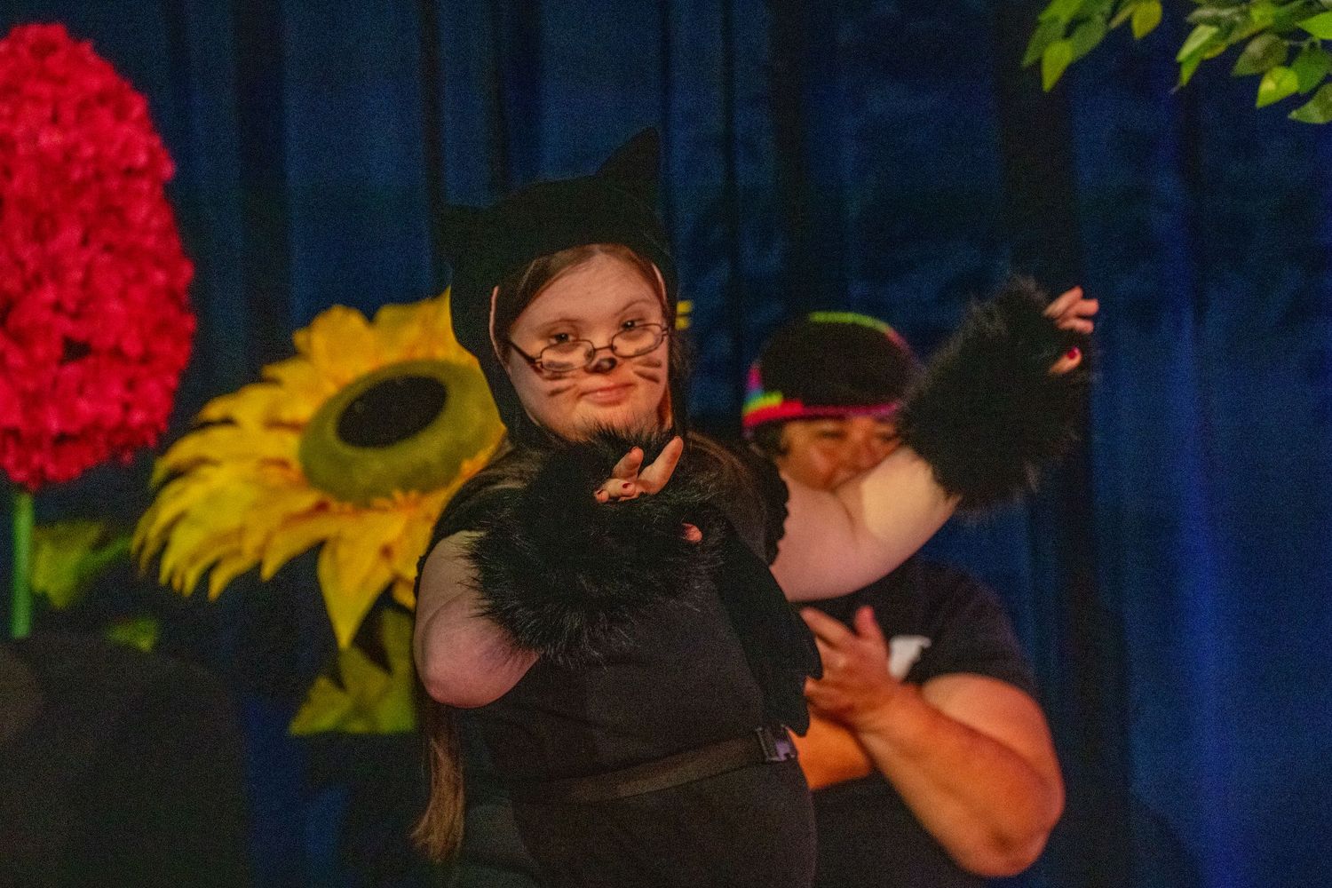 Performer in black cat costume dances on stage with colorful flower backdrop during theatrical production.