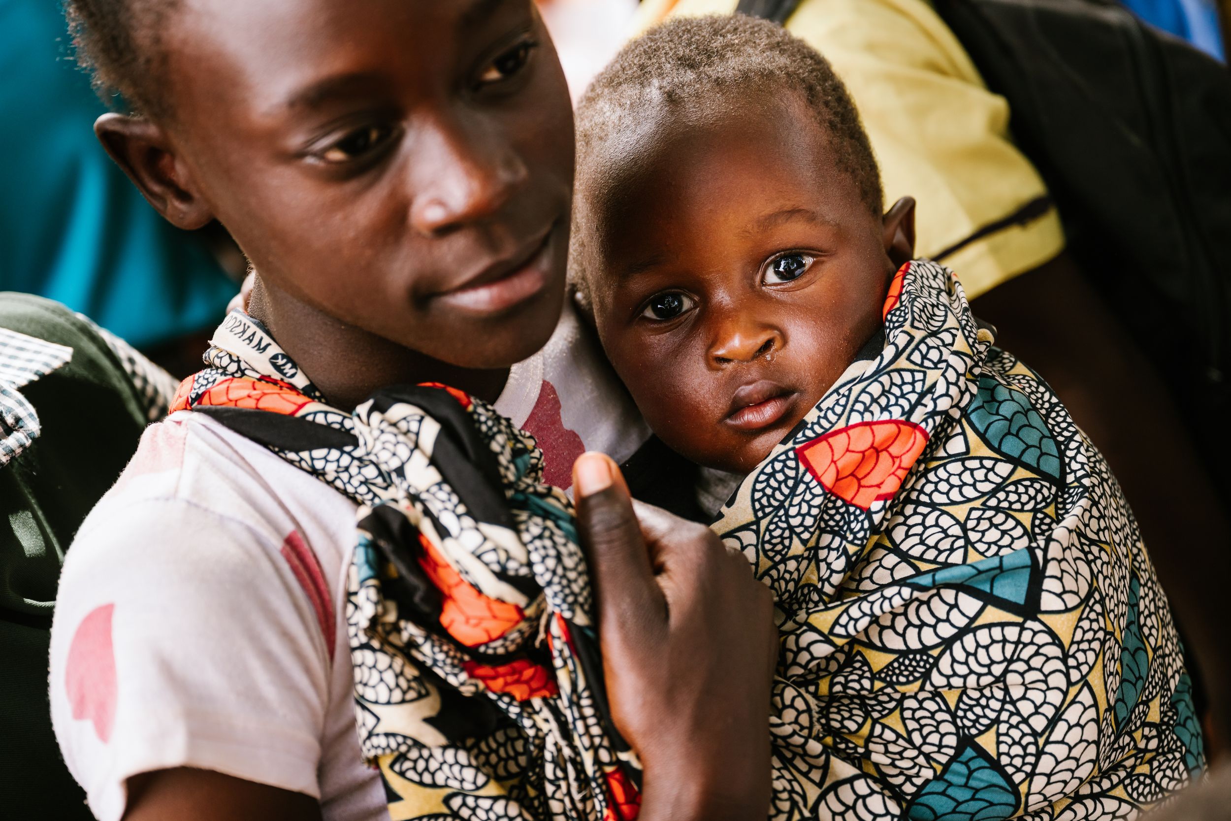 Young African child held by an older sibling, both wearing colorful traditional fabric clothing in a candid portrait.