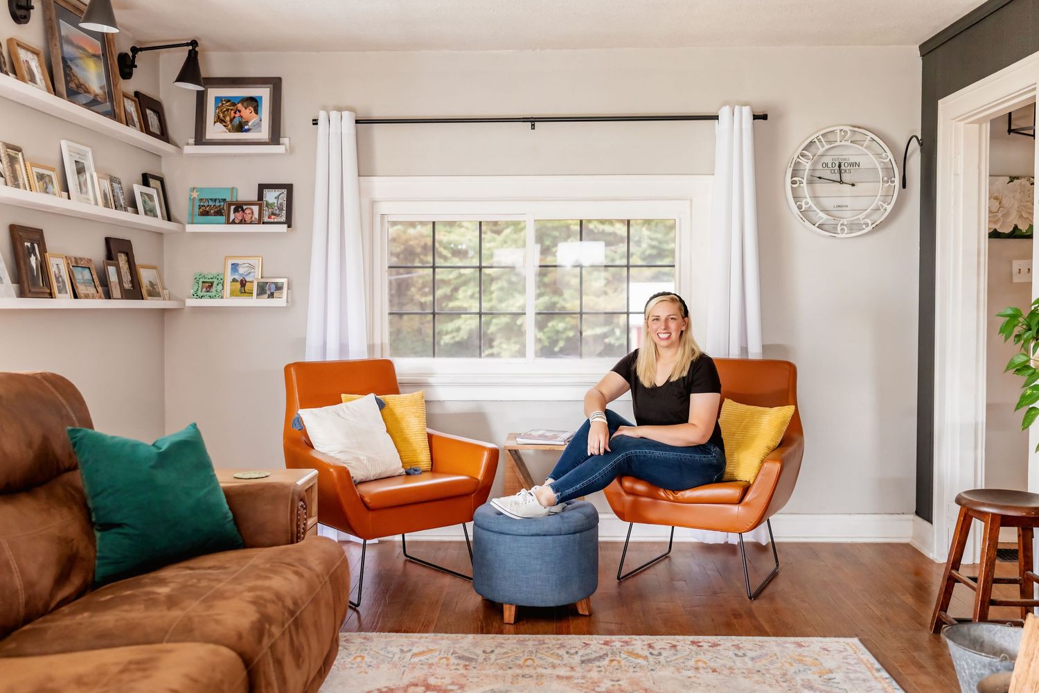 Woman sitting between two colorful accent chairs in a bright living room with built-in shelves and hardwood floors.