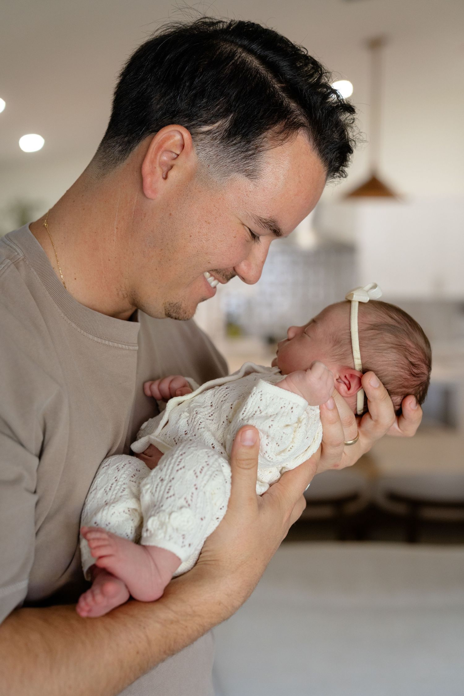 A smiling father tenderly holds his newborn baby in a white floral outfit with a bow headband indoors.