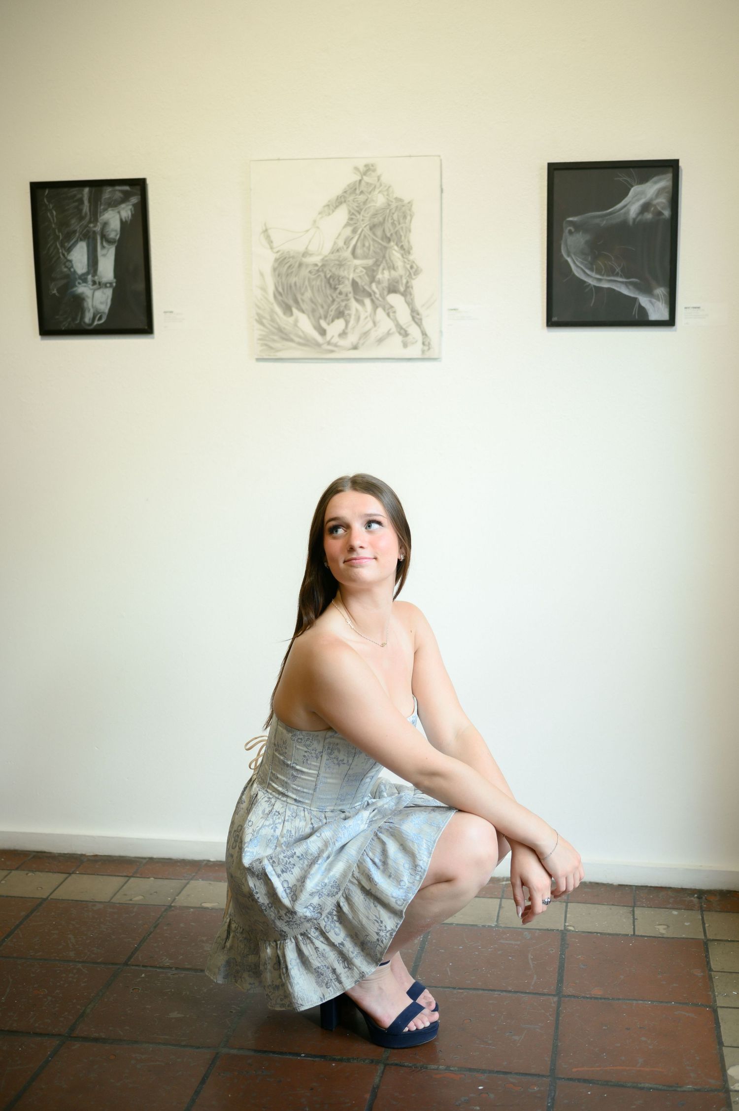 Young woman in gray dress crouching on tile floor in art gallery with three framed drawings on white wall behind her.