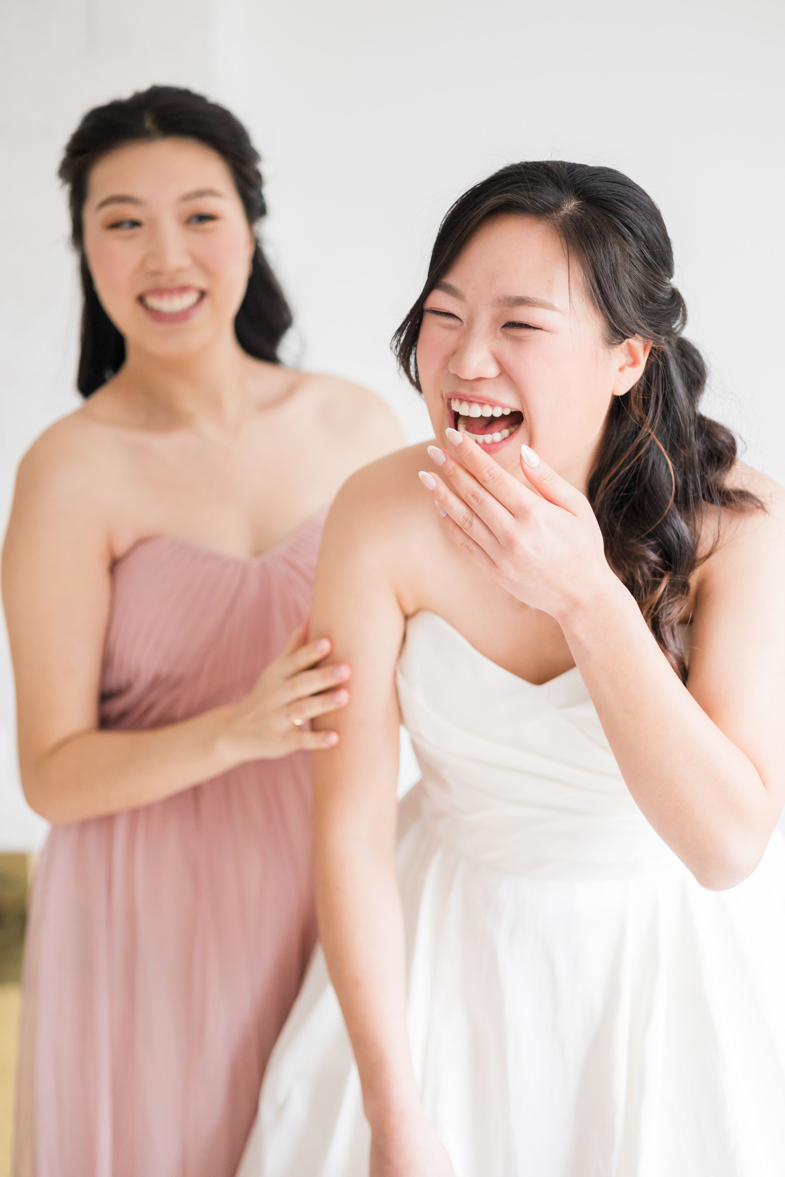 Bride in white gown laughs joyfully as bridesmaid in pink dress helps her get ready on wedding day.