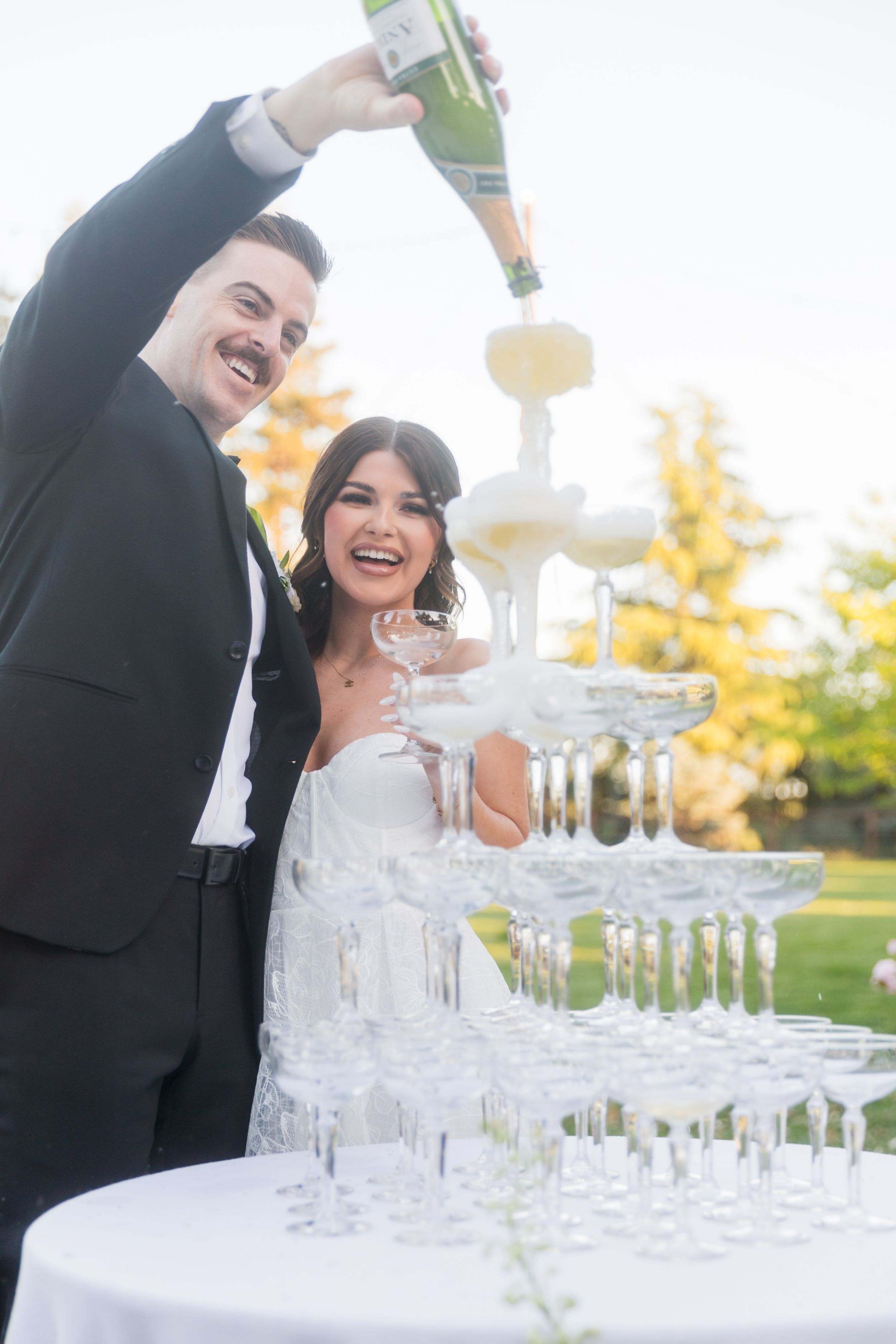 Bride and groom laughing while pouring champagne tower at outdoor wedding reception.