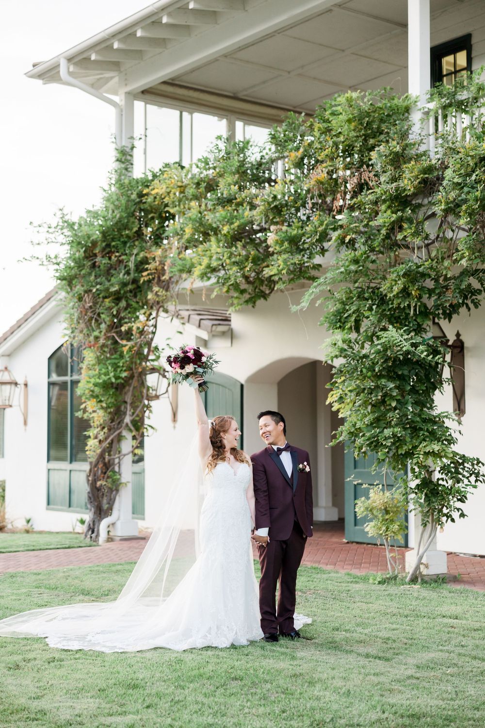 Bride raises bouquet while groom smiles beside her in front of a vine-covered estate on their wedding day.