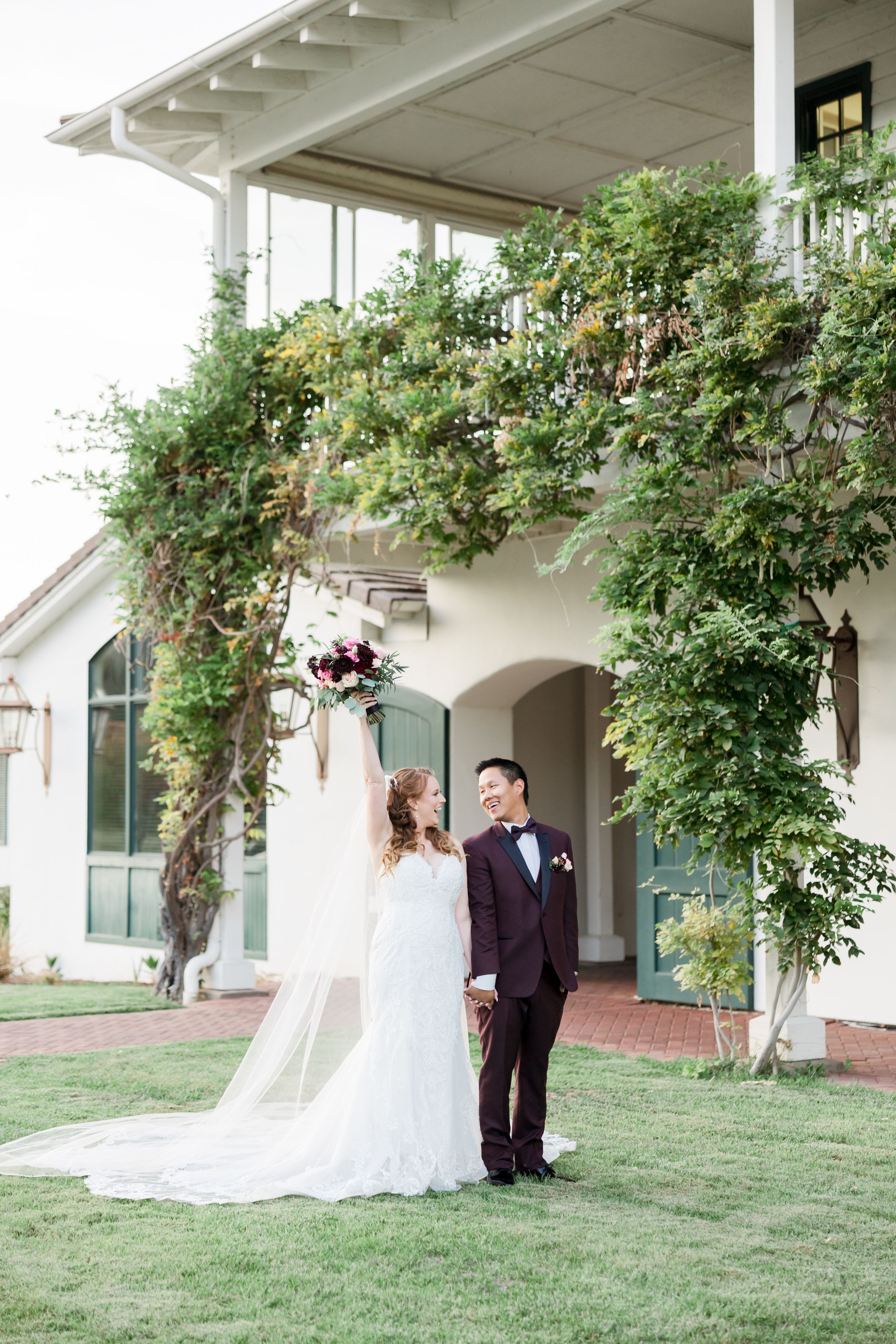 Bride raises bouquet while groom smiles beside her in front of a vine-covered estate on their wedding day.