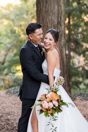 Bride and groom embracing in a redwood forest, bride holding a colorful bouquet of orange and pink flowers in a white gown.