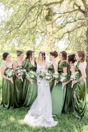 Bride in white gown with bridesmaids in green satin dresses holding white bouquets outdoors under oak trees.
