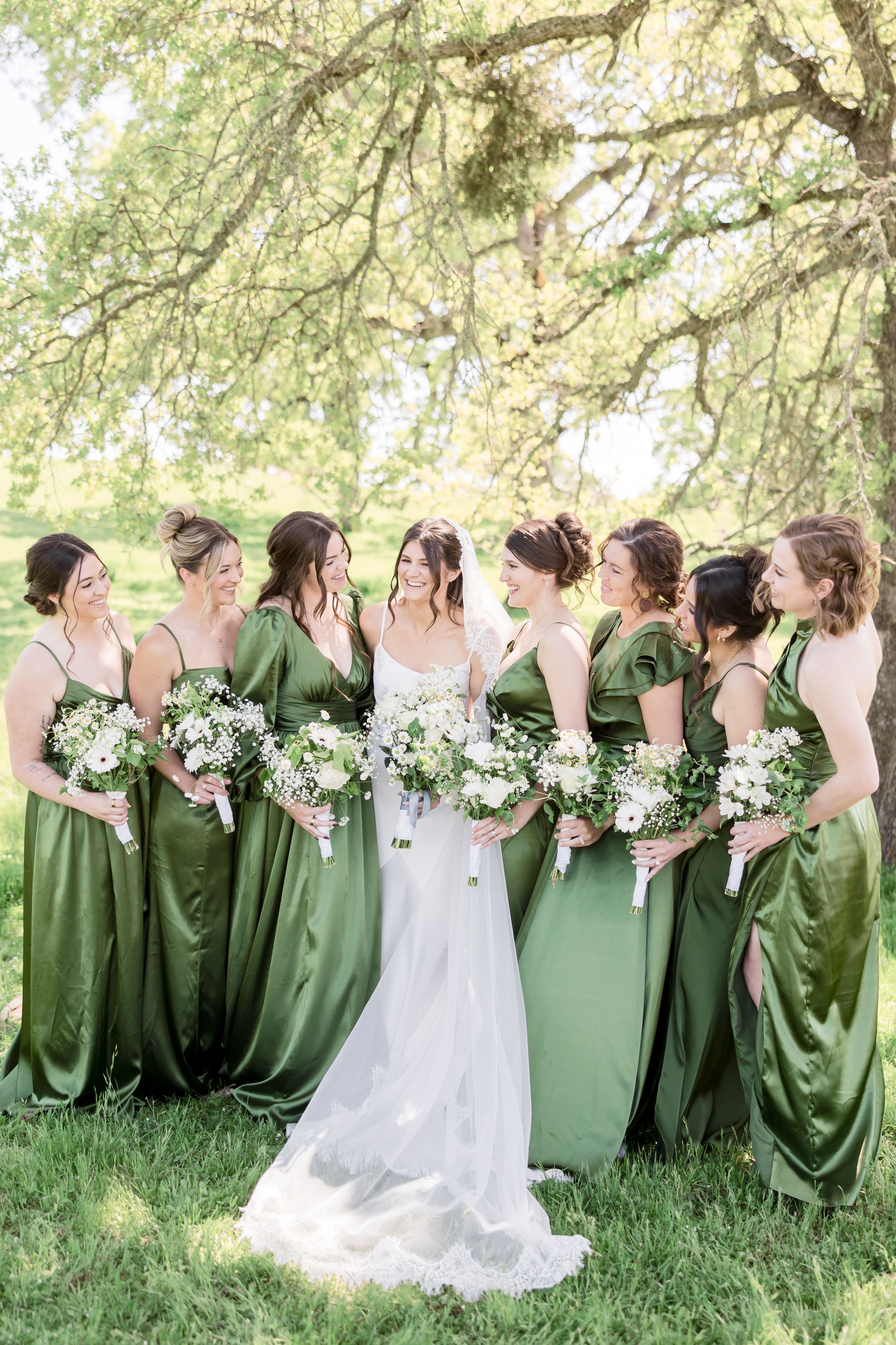 Bride in white gown with bridesmaids in green satin dresses holding white bouquets outdoors under oak trees.