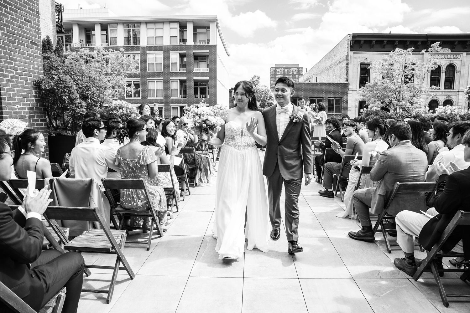 Black and white photo of a bride and groom walking down the aisle at an outdoor rooftop wedding ceremony.
