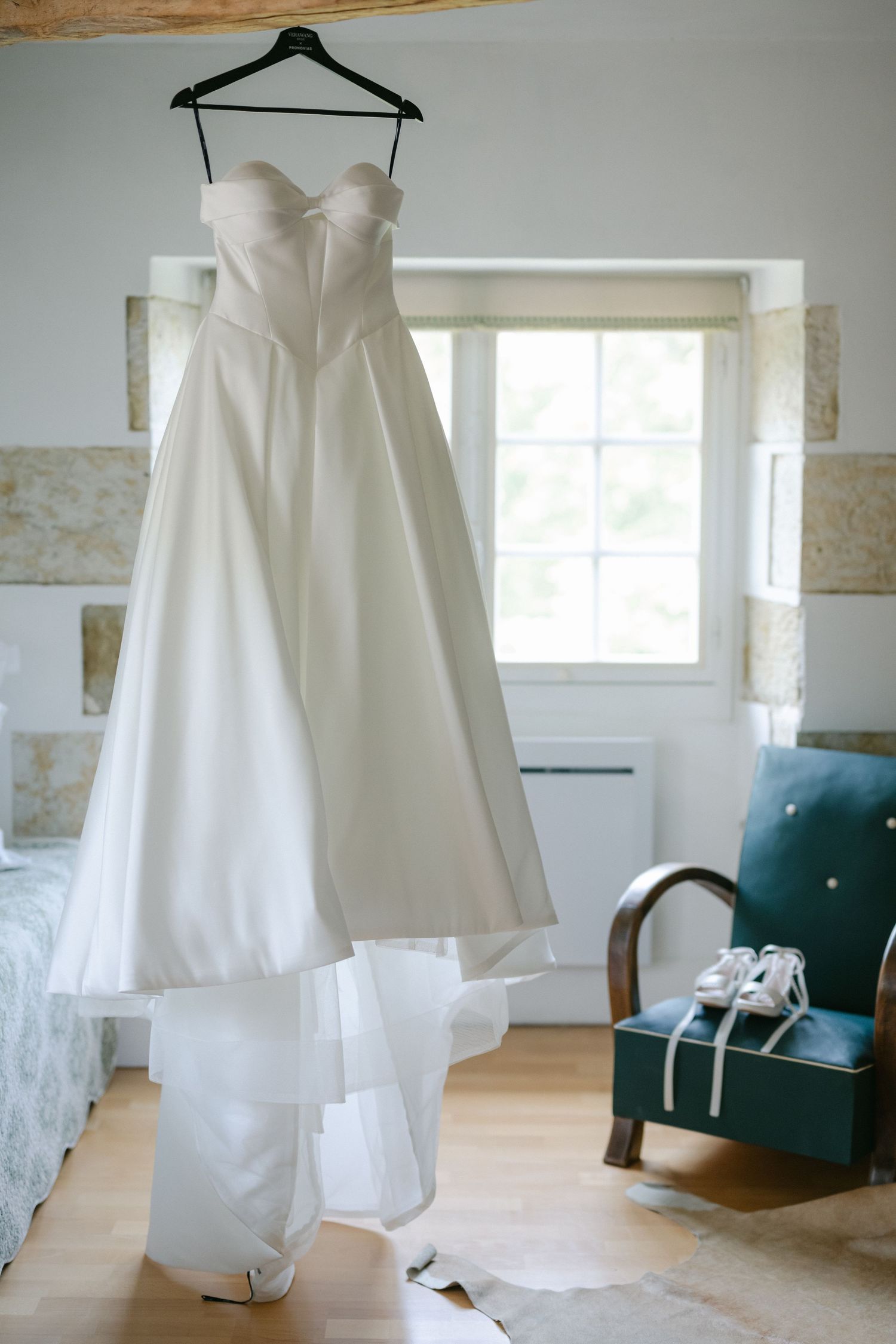 White bridal heeled sandals with ribbon laces displayed on a teal velvet armchair in an elegant wedding venue.