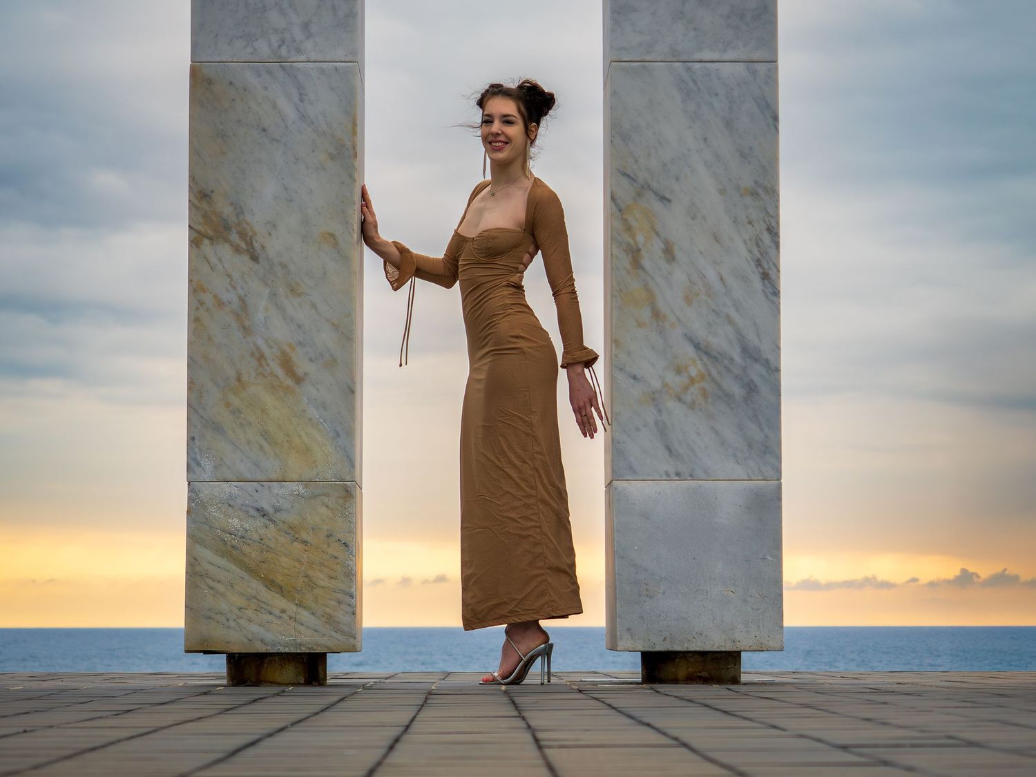 Woman in elegant tan maxi dress poses between marble columns at sunset overlooking the ocean.