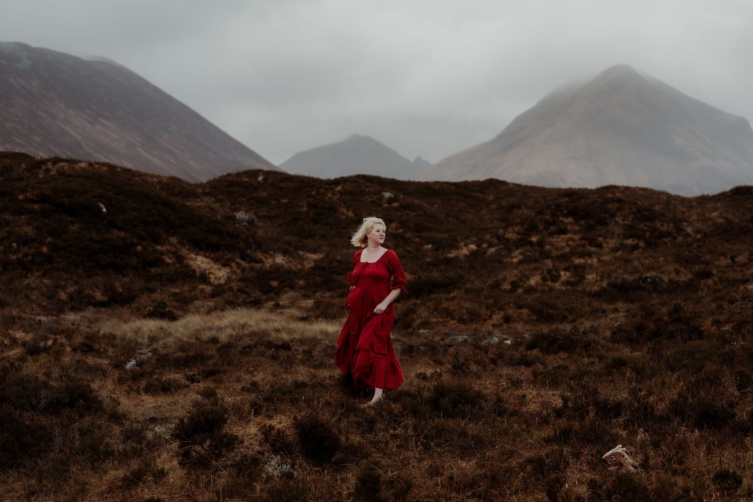 Woman in red dress walking through misty Scottish Highland moorland with dramatic mountains in background.
