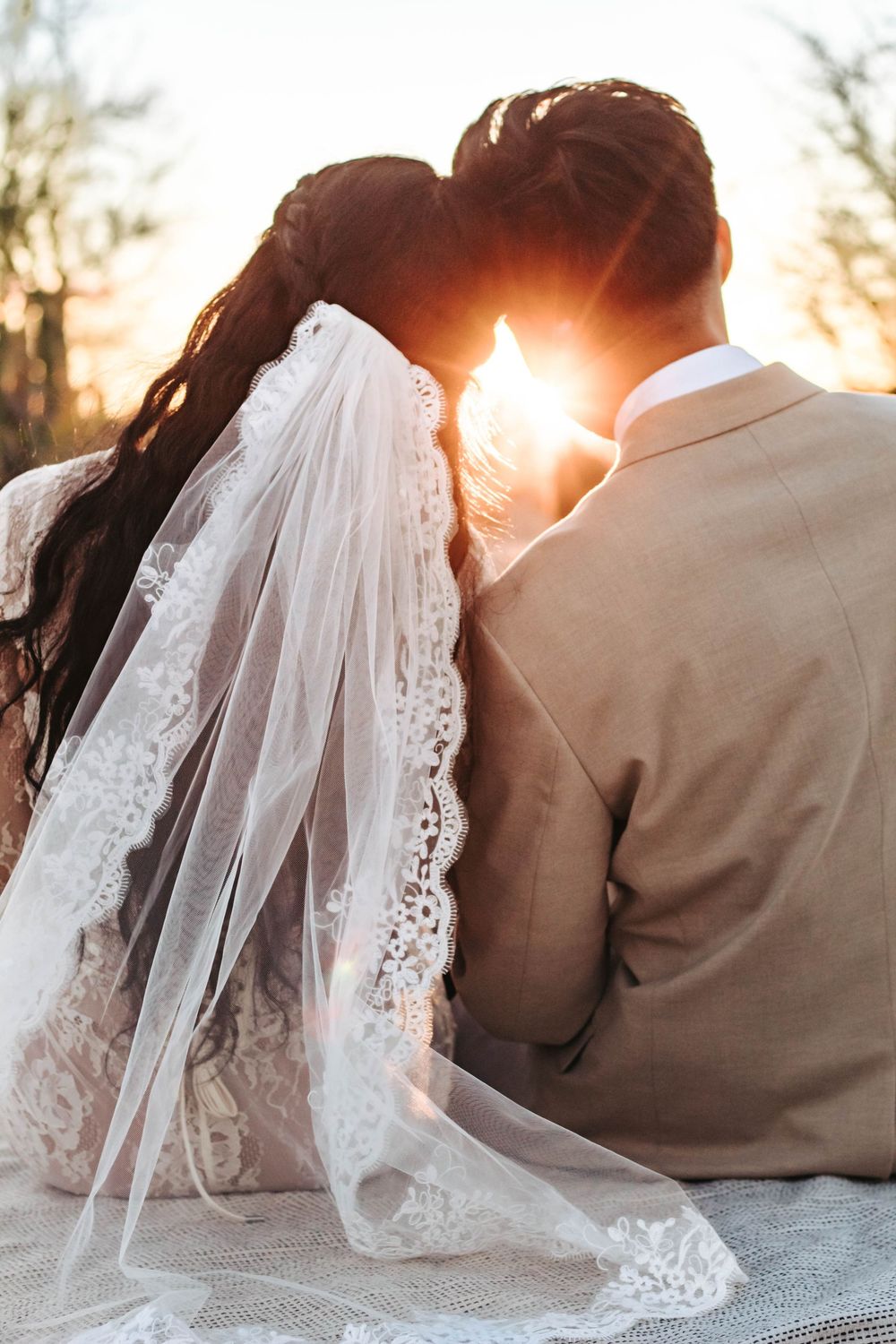 Bride and groom lean on each other looking at sunset as it peeks from between their heads.