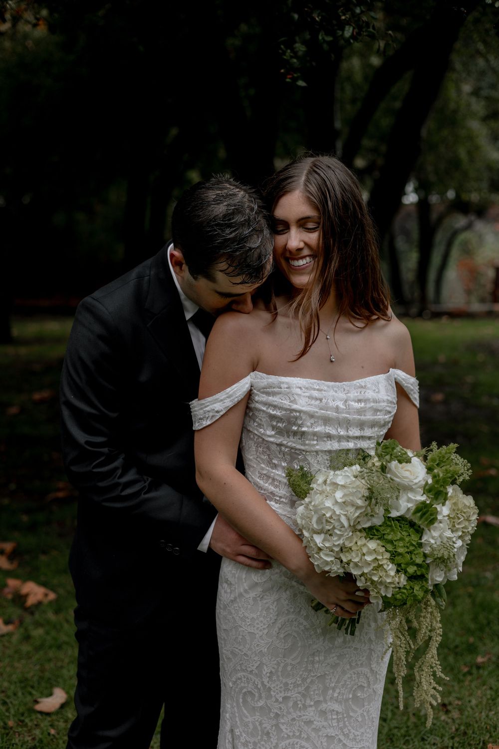 Groom kisses brides shoulder while she smiles at camera.