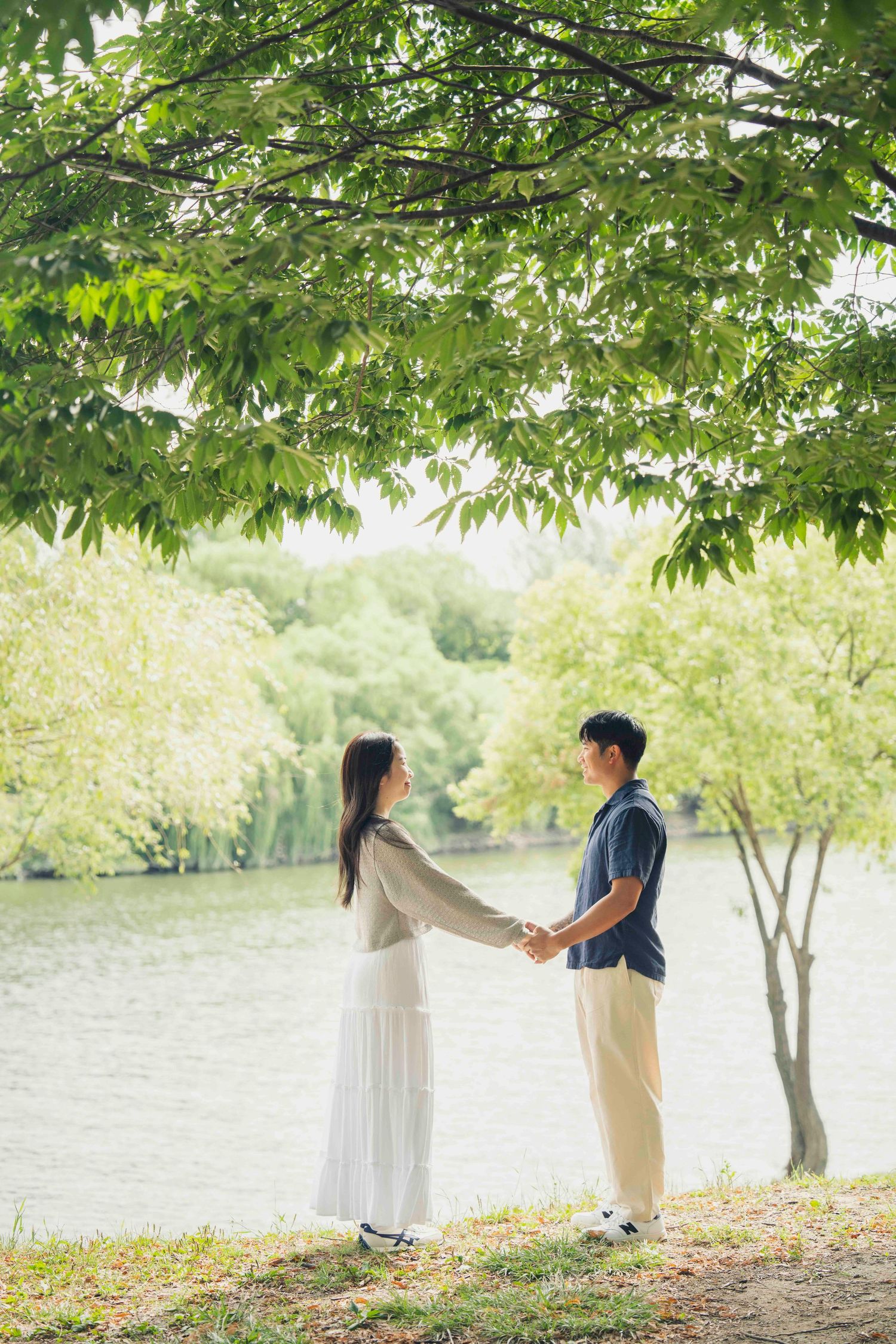 A couple holds hands facing each other near a serene lake surrounded by lush green trees in a park.