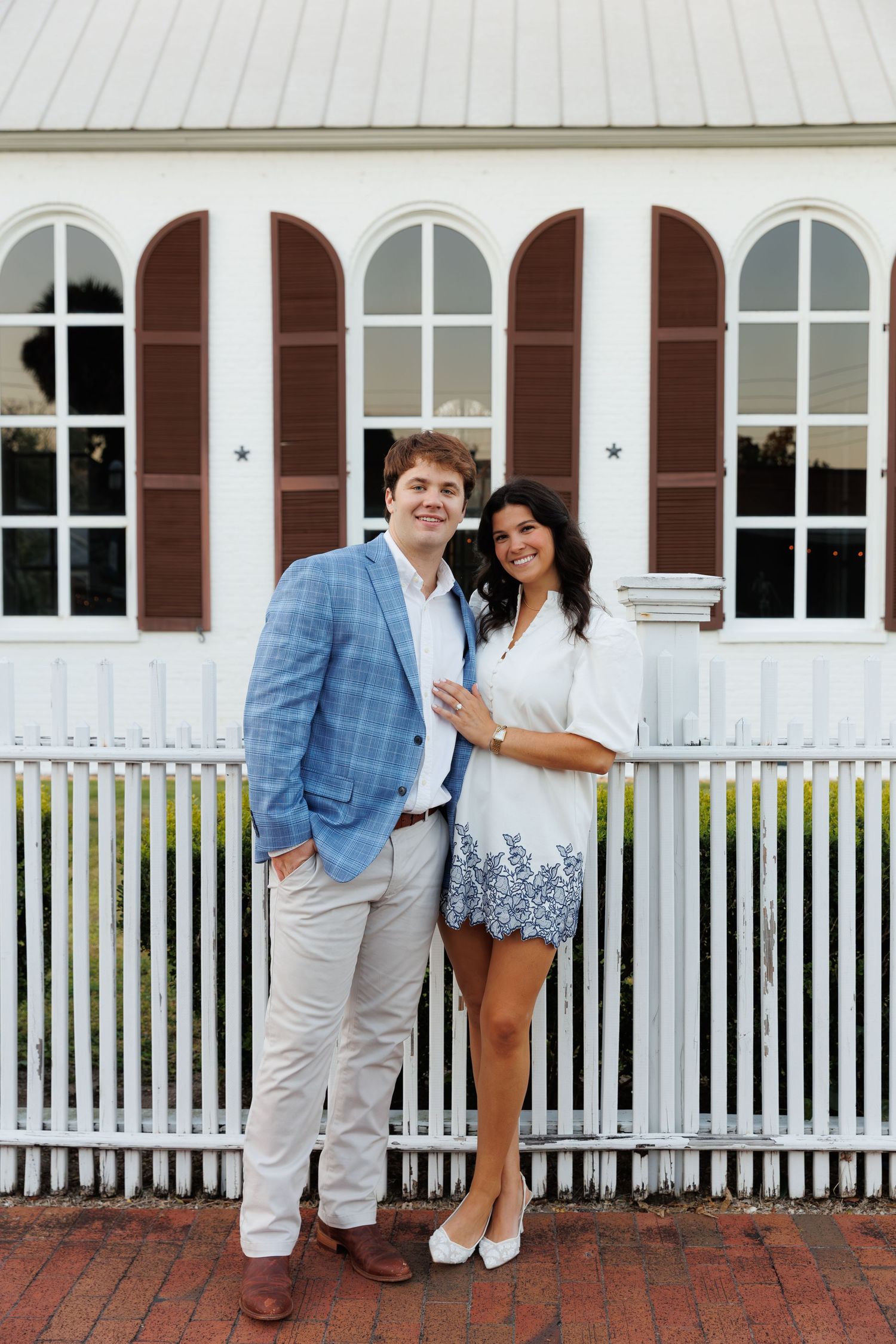 Red-haired woman in camel sweater and dark shorts standing by white picket fence outside historic building.
