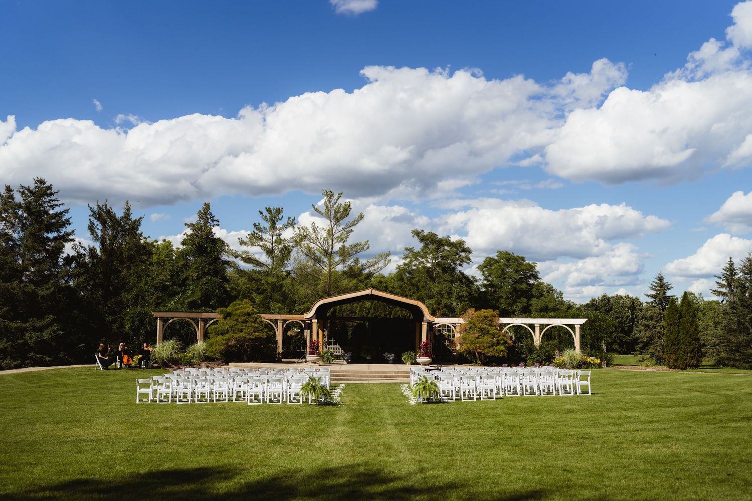 Outdoor wedding ceremony setup with white chairs on green lawn, rustic barn backdrop, and blue sky with clouds.