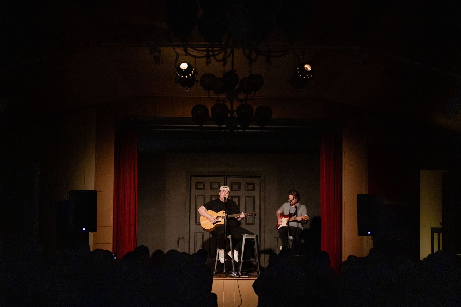 Two musicians perform acoustic guitars on a dimly lit stage with red curtains during an intimate live concert.