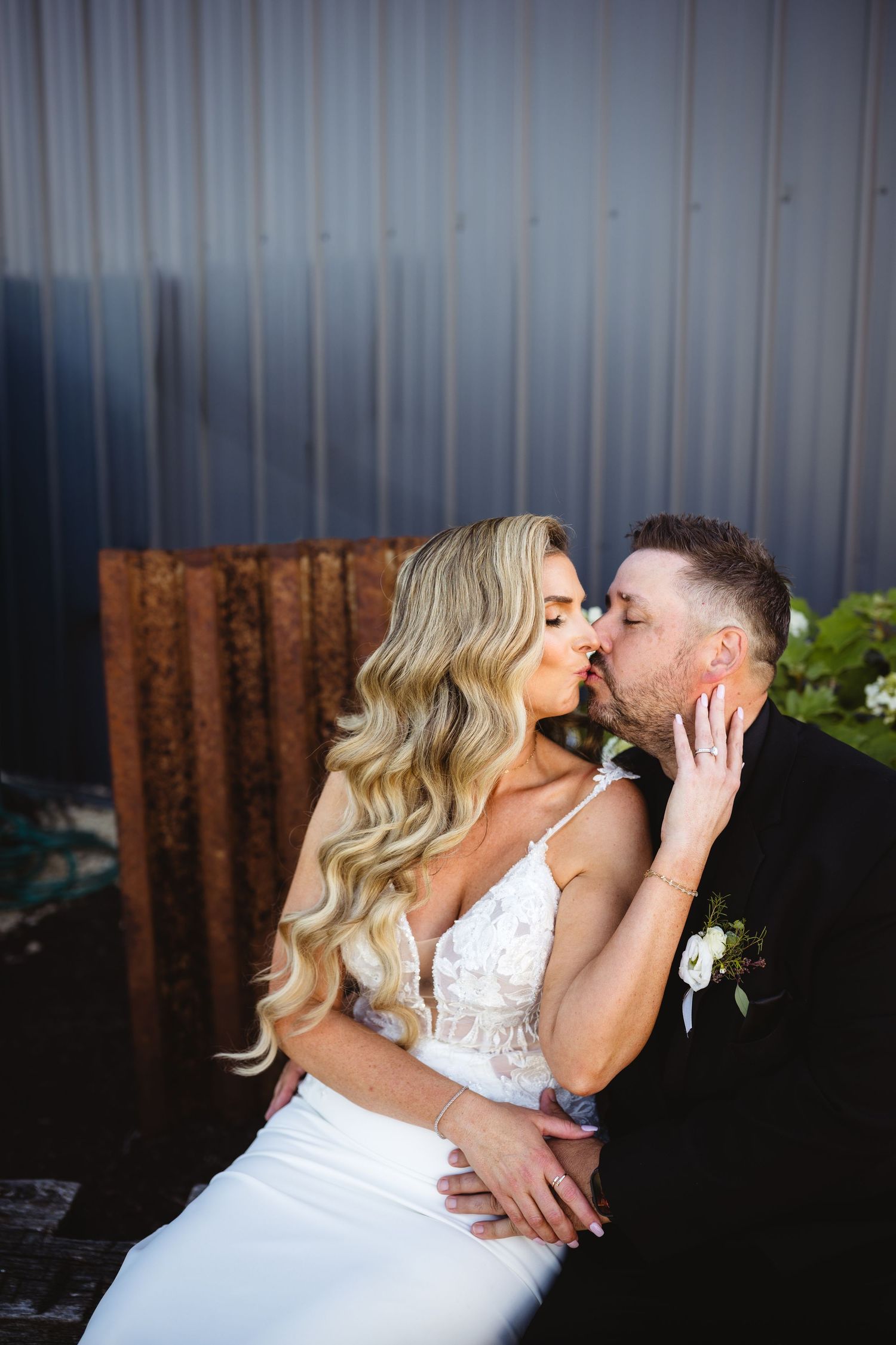 A bride and groom share a romantic kiss while seated outdoors, the bride in a white lace gown with long wavy blonde hair.