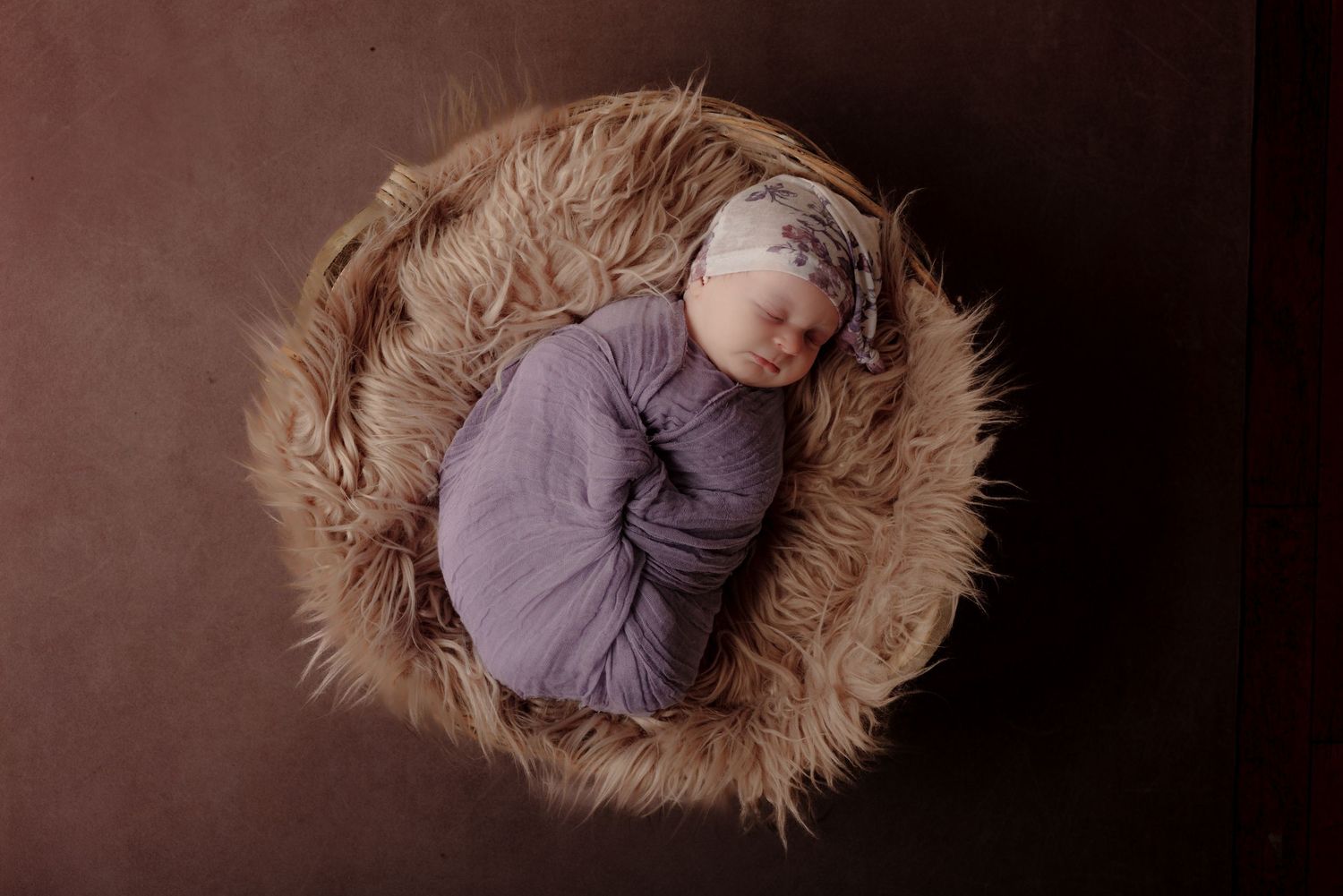 Newborn baby swaddled in purple wrap with floral headband sleeping in a furry brown bowl prop.