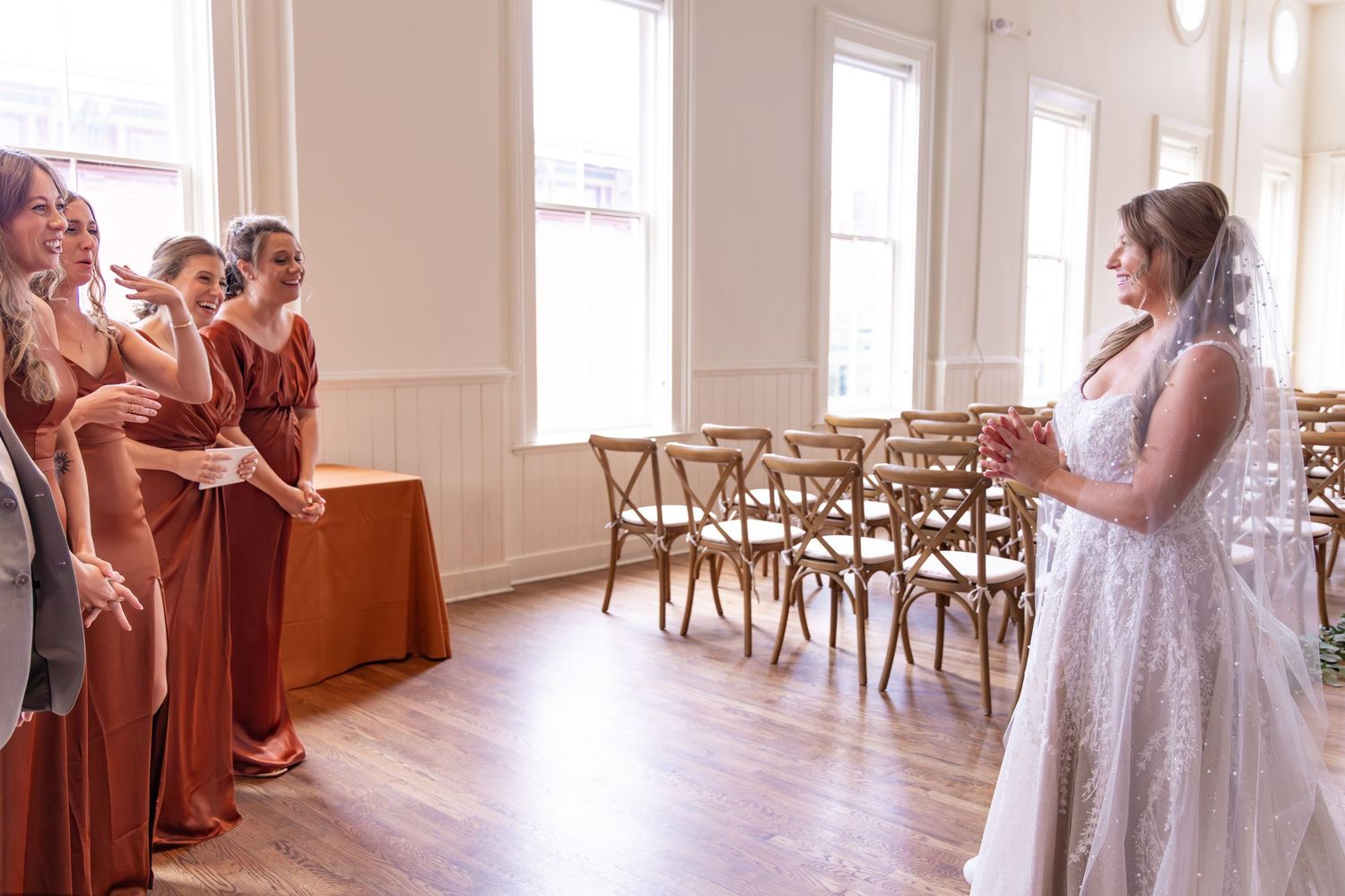 Bride in white lace gown reveals her look to bridesmaids in rust-colored dresses inside elegant venue.