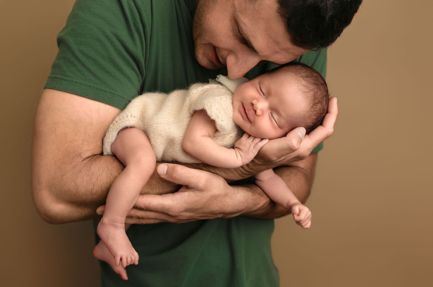 Dad cradles his new baby daughter, as she smiles in his arms against a rich tan backdrop, by Charlottesville newborn photographer.