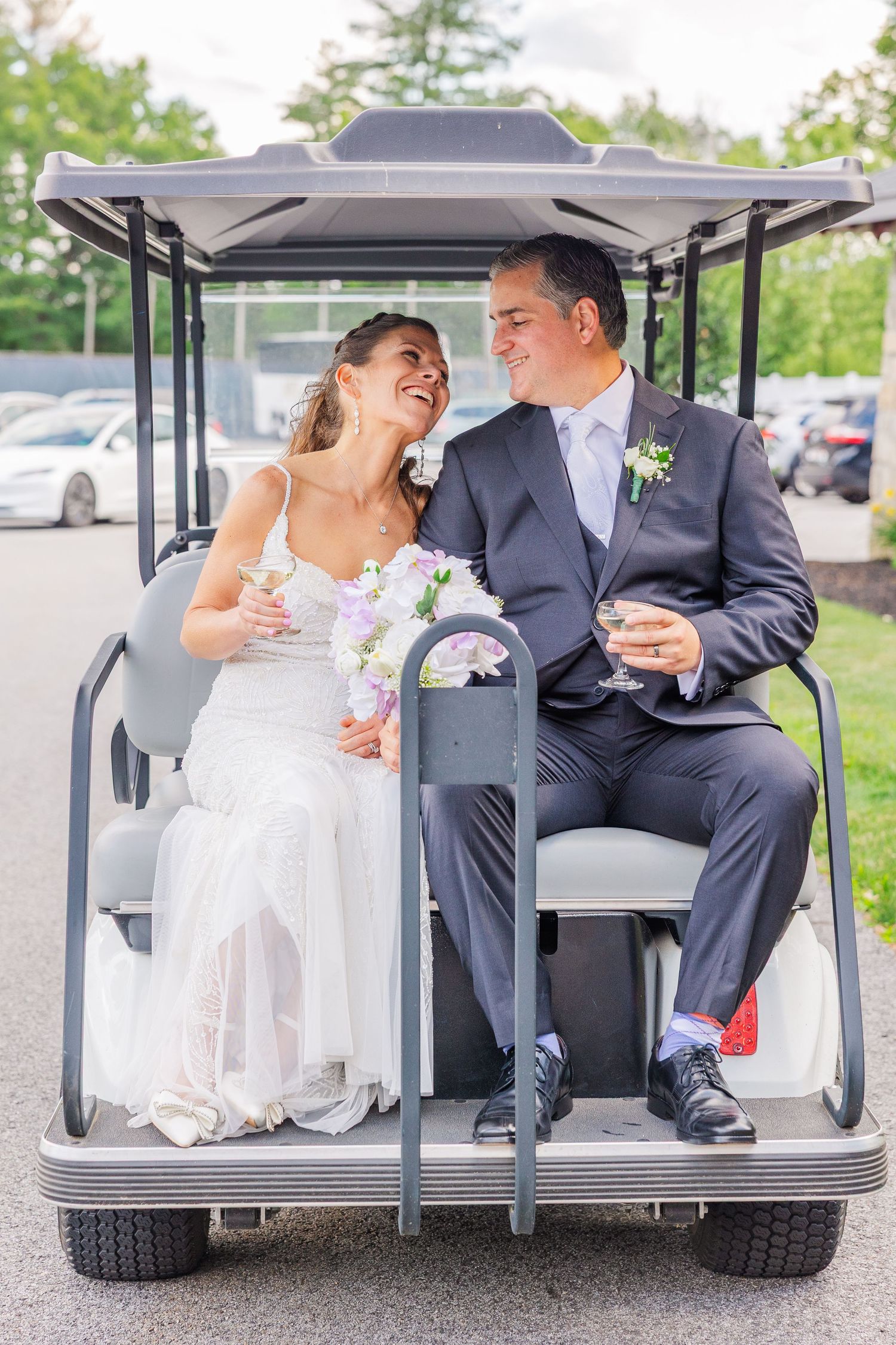 Bride and groom laughing together on a golf cart, holding champagne glasses and a bouquet on their wedding day.