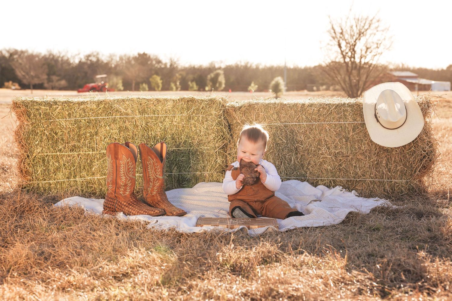 Baby sits on blanket by hay bales with cowboy boots and hat in golden sunlit farm field.