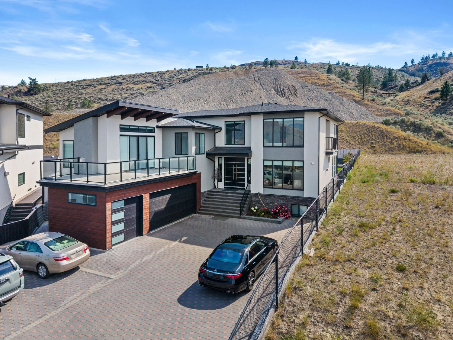 Aerial view of a modern two-story home with large windows, double garage, and spacious driveway against a hillside backdrop.